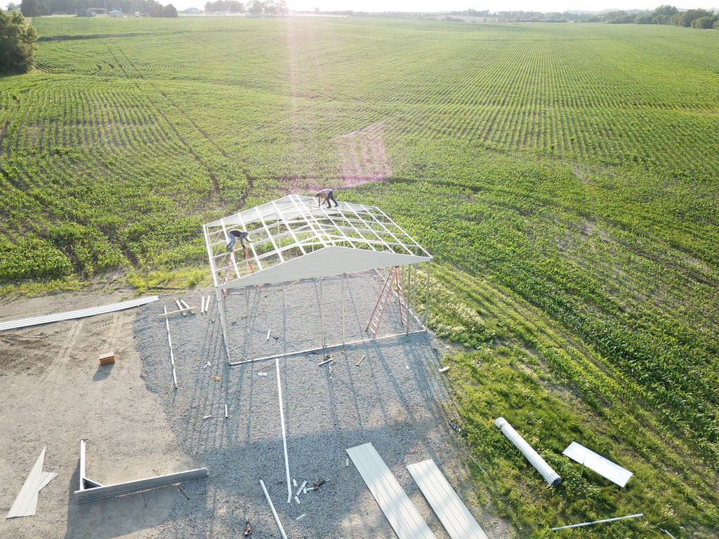Aerial photo of a building by NESheds in the process of being built. Roofers put the main support beams into place. Cornfields spread the course of the background.